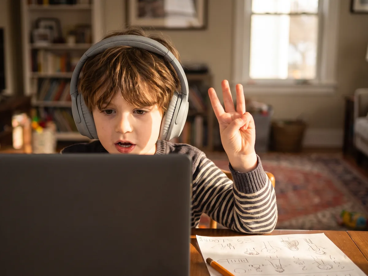 Child in headphones answering out loud during a live one-on-one online math lesson, holding up three fingers