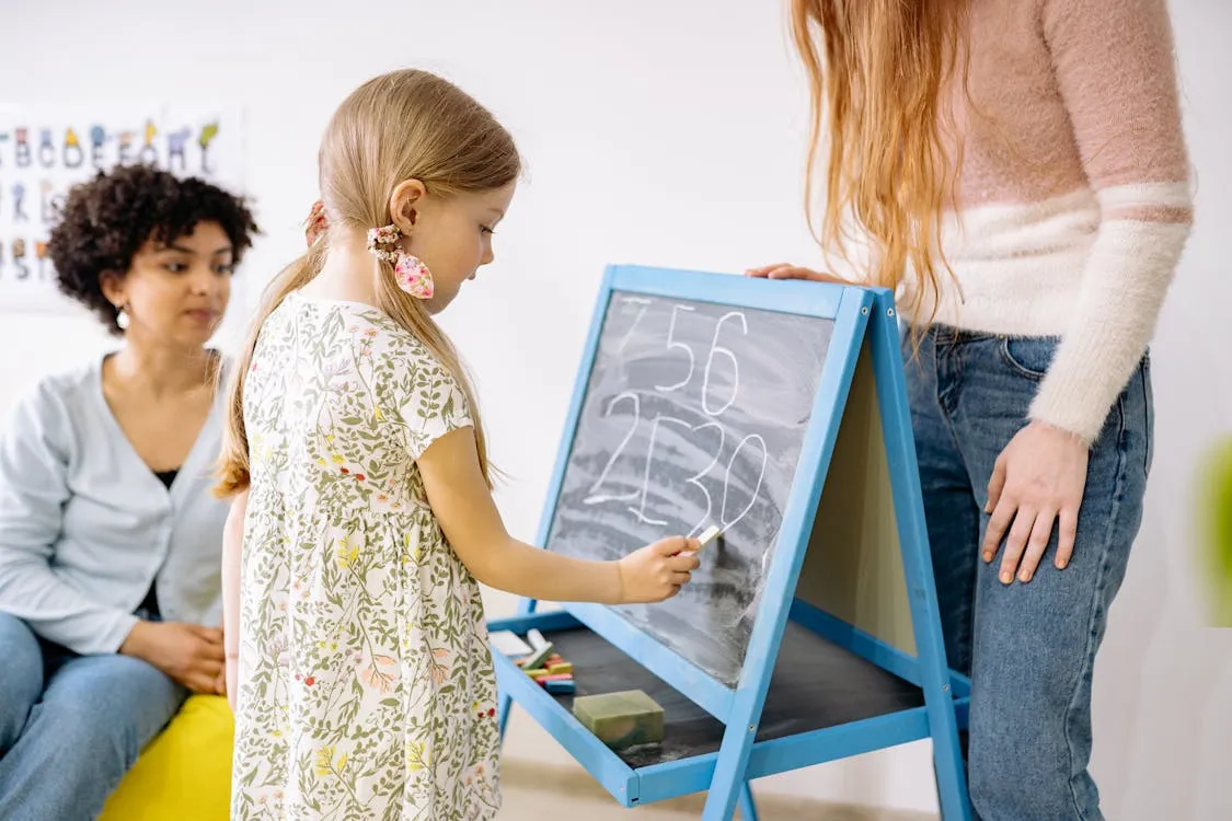 Young student writing math numbers on a chalkboard with teacher